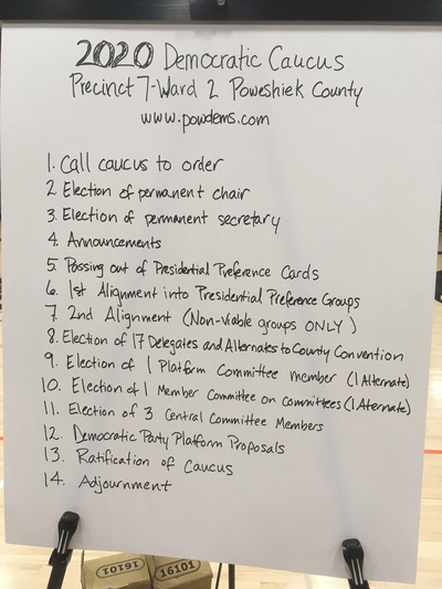 A hand-written sign on an easel.  2020 Democratic Caucus.  Precinct 7-Ward 2 Poweshiek Count.  www.powdems.com.  1. Call caucus to order.  2. Election of permanent chair.  3. Election of permanent secretary.  4. Announcements.  5. Passing out of Presidential Preference Cards.  6. 1st Alignment into Presidential Preference Groups.  7. 2nd Alignment (Non-viable groups ONLY).  8. Election of 17 Delegates and Alternates to County Convention.  9. Election of 1 Platform Committee member (1 Alternate).  10. Election of 1 Member Committee on Committees (1 Alternate).  11. Election of 3 Central Committee Members.  12. Democratic Party Platform Proposals.  13. Ratification of Caucus.  14. Adjournment.