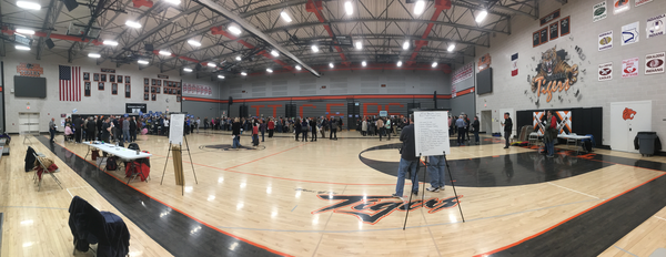 A panoramic shot of a gym.  In the foreground are two easels with the rules.  People are scattered in groups, with a small group in the center, and some folks by themselves at the right.