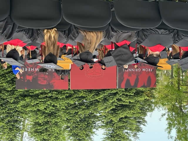 A picture of the commencement stage.  At the left is a large panel that says 'From Grinnell', superimposed on a red and black picture of somewhere on campus.  In the middle is a red panel with the Grinnell emblem.  At the right is the phrase 'to everywhere', superimposed on some faces in red and black.  We can see people on stage, students in the foreground, and some trees in the background.