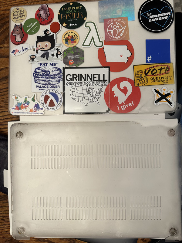 A picture of the two halves of a MacBook cover. The top half has a lot of stickers on it as well as a broken piece in the lower-right-hand corner, held on only by a 'no kings' sticker. The feet are gone from the bottom and there's a big chunk missing from the upper-right corner.