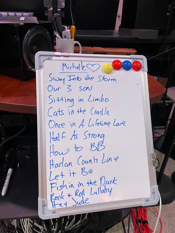 A whiteboard with 'Michelle' and a heart at the top along with the songs 'Sway Into the Storm', 'Our 3 sons', 'Sitting in Limbo', 'Cats in the Cradle', 'Once in A Lifetime Love', 'Half As Strong', 'How to BB', 'Harlan County Line', 'Let it Be', 'Fishin in the Dark', 'Rock + Roll Lullaby', and 'Hey Jude'