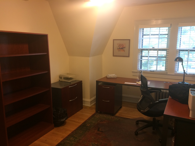 An office with two windows,
a large L-shaped desk, two file cabinets, a desk lamp, and a set of
empty bookshelves.