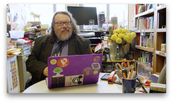 Sam Rebelsky, seated in his very messy office.  Sam's purple laptop is in front of him.  Sam is wearing a natty blue tie with geometric shapes.  Behind Sam are way too many piles of paper.  On the table in front of Sam is a wine glass, a jar full of pens, and a stapler.