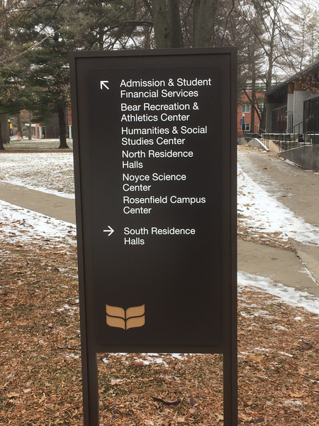 A photo with a sign that has an arrow pointing diagonally to the left and six entries: 'Admission & Student Financial Services', 'Bear Recreation & Athletics Center', 'Humanities & Social Studies Center', 'North Residence Halls', 'Noyce Science Center', 'Rosenfield Campus Center'.  There is also an arrow pointing to the right with 'South Residence Halls'.  The Forum is visible to the right and Noyce is visible straight ahead.