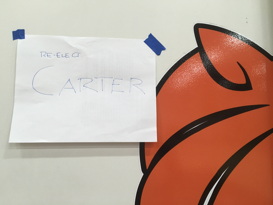 A piece of 8.5x11 paper taped to the wall with blue tape, next to the school logo.  On the paper is written 'Re-elect CARTER'.