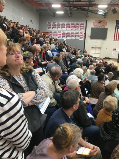 A large number of people, sitting in the stands of a gym.  You can see white pieces of paper in the hands of some.  In the background, you see championship banners for the Grinnell tigers and a part of an American flag.